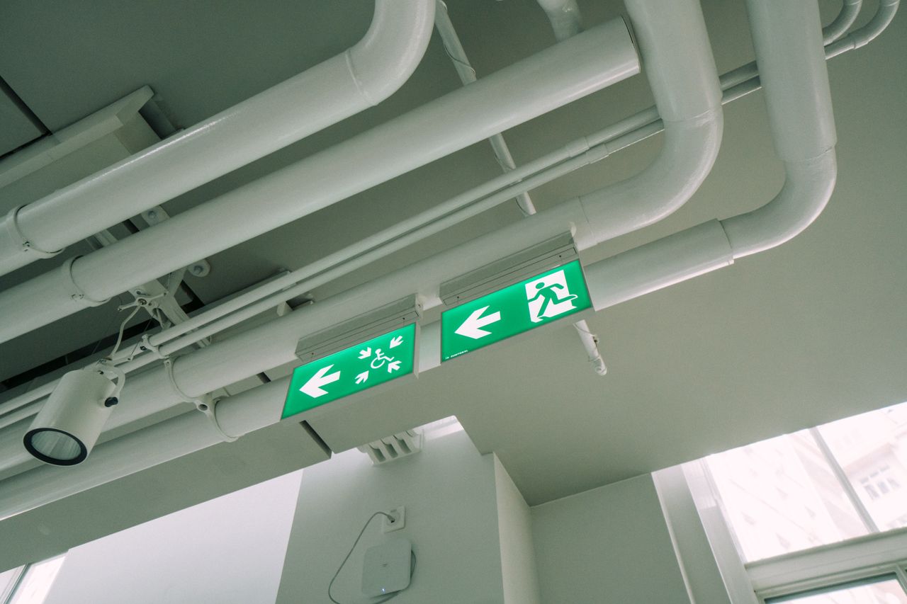White ceiling with industrial pipes and green exit signs in a modern building.